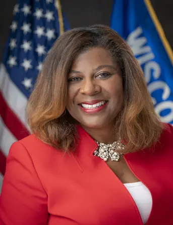 Official portrait of State Senator LaTonya Johnson, a Wisconsin Democrat, smiling warmly. She is wearing a bright red dress and standing in front of the Wisconsin state flag. Her hair is styled in loose waves, and the portrait highlights her commitment to public service and leadership in the state legislature.