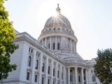 The Wisconsin state capitol on a sunny day. The sky is clear and the sun is shining from behind the dome. There are green trees in the foreground.