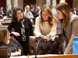 State Representative Dianne Hesselbein speaks to other legislators from Dane County on the floor of the assembly. They are all white woman with dark hair and are leaned over a single desk, discussing something together.