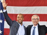 Governor Tony Evers and Lt. Governor Mandela Barnes stand together in front of an oversized American flag. Gov. Evers is an older white man with glasses wearing a suit and blue tie. Lt. Gov. Barnes is a Black man with a shaved hear wearing a suit and raising his hand to wave at someone.