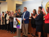 State Senator Tim Carpenter stands behind a lectern at a press conference. He is a tall man with reddish brown hair and glasses wearing a blue blazer. On the front of the lectern is a large sign with the silhouette of WI colored with the Progress Pride flag  and black text that reads "2025 Equality Agenda". Senator Carpenter is joined by at least 14 of his legislative colleagues who are clapping as he speaks.