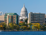 The Wisconsin State Capitol as seen from on or across a lake. The sky is blue and other modern city bulidings can bee seen beside it.