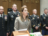State Representative Dianne Hesselbein speaks on the Assembly floor standing in fornt of at least five military members. She is a white woman with medium brown hair wearing a grey blazer speaking into a microphone attached to the lectern in front of her.