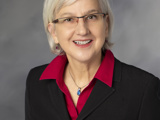 The official photo for WDVA Secretary Mary Kolar. She is an older white woman with short white hair and glasses. Behind her is a neutral grey background.
