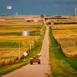 An image of a farm vehicle driving on a rural road between corn fields