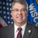 A formal portrait of Senator Brad Pfaff. He is a middle-aged white man with glasses and dark hair. He is wearing a dark suit with a white shirt and a red tie. In the background are the flags of the United States and the state of Wisconsin.