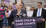 Senator Kelda Roys stands at a lectern speaking into a cluster of microphones at a press event. She is a white woman with blonde hair pulled back. On the front of the lectern is a purple sign with light pink text that reads, "Save Child Care Counts" and a hashtag showing the same below it. There are many people gathered behind her with various handmade protest signs, including other legislators. They are outside and the weather seems nice.