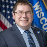 A formal portrait of Senator Mark Spreitzer. He is a white man with glasses and dark hair. He is wearing a dark suit with a light blue shirt and a tie with a blue geometric pattern. In the background are the flags of the United States and the state of Wisconsin.