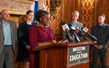 Senator LaTonya Johnson stands behind a lectern at a press conference. She is a Black woman with her hair pulled back. She is wearing a red dress and looking out at the audience as she speaks into a cluster of microphones on the lectern. On the front of the lectern is a dark sign with white text that reads, "Investing in our kids' education." Behind her, at least four people can be seen gathered in support.