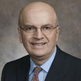 A formal portrait of Senator Bob Wirch. He is a white man with a bald head and thin glasses. He is wearing a dark suit with a light blue shirt and a brown tie. The background of the photo is a grey/brown abstract backdrop.