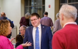 Senator Mark Spreitzer speaks to two people at a swearing-in event. Senator Spreitzer is a white man wearing glasses and a blue suit and tie. He is speaking to a woman with curl yhair and a pink shirt. Also in the conversation is an older man with white hair wearing a red shirt. Both the other people are facing away from the camera. In the background, many other people appear to be mingling.