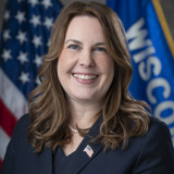 A formal portrait of Senator Dianne Hesselbein. She is a white woman with blue eyes and brown hair. She is wearing a blue blazer and smiling for the camera. In the background are the flags of the United States and the state of Wisconsin.