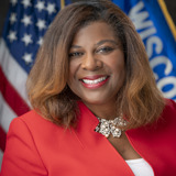 A formal portrait of Senator LaTonya Johnson. She is a Black woman with thick brown hair down to her shoulders. She is wearing a white shirt and a bright red jacket that matches her red lipstick. In the background are the flags of the United States and the state of Wisconsin.