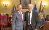 Senator Brad Pfaff stands beside Governor Tony Evers for a photo. Senator Pfaff is a middle-aged white man wearing a light grey suit, glasses, and a red tie. Governor Evers is an older man with white hair and glasses wearing a dark wuit with a green tie. In the background are the flags of the United States, the state of Wisconsin, and the Wisconsin National Guard.