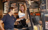 Senator Dianne Hesselbein sits in the driver's seat of a forklift and grins at the man standing beside her. The man is wearing a dark t-shirt and jeans and pointing to a clipboard that's hanging in the forklift.