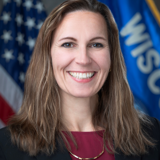 A formal portrait of Senator Melissa Ratcliff. She is a white woman with long brown hair. She is wearing a dark blazer over a berry red shirt. In the background are the flags of the United States and the state of Wisconsin.