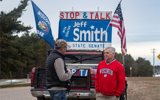 Senator Jeff Smith speaks to a constituent standing on the street. Senator smith is wearing a red jacket and listening to a man in a lback vest and camo hat. Behind them is Senaotr Smith's truck with a large sign that says "Stop and Talk with Senator Jeff Smith." There are two full-sized flags flying from the truck - one for the state of Wisconsin and one for the United States.