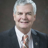 A formal portrait of Senator Jeff Smith. He is a white man with greying white hair. He is wearing a dark suit with a white shirt and a brown striped tie. The background of the photo is a grey/brown abstract backdrop.