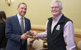 Senators Chris Larson and Jeff Smith hold a gavel together and pose for the camera. Senator Larson has short brown hair and a beard and is wearing a blue suit. Senator Smith is slightly older with white hair and glasses. He is wearing a pink shirt with a black Wisconsin State Senate vest over it.