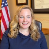 A formal portrait of Senator Kelda Roys. She is a white woman with medium length blonde hair and blue eyes. She is wearing a blue sweater and a pearl necklace. Behind her are two framed documents handing on the wall and an American flag. 