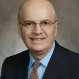 A formal portrait of Senator Bob Wirch. He is a white man with a bald head and thin glasses. He is wearing a dark suit with a light blue shirt and a brown tie. The background of the photo is a grey/brown abstract backdrop.