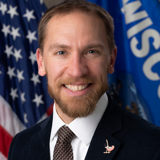 A formal portrait of Senator Chris Larson. He is a white man with brown hair and a beard. He is wearing a dark suit with a white shirt and a dark patterned tie. In the background are the flags of the United States and the state of Wisconsin.