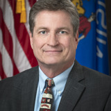 A formal portrait of Senator Tim Carpenter. He is a middle-aged white man with blue eyes and dark hair. He is wearing a dark suit with a light blue shirt and a tie with various logos for the University of Wisconsin on it. In the background are the flags of the United States and the state of Wisconsin.