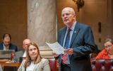 Senator Bob Wirch stands on the floor of the Wisconsin State Senate giving a speech. He is an older white man with a bald head wearing a suit and a tie with the American flag on it. He is holding a piece of paper. Other senators are seated nearby, looking up at him and listening to him speak.