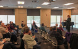 Senator Tim Carpenter speaks to a room full of people. He is standing at the front of the room, which is on the right side of the photo. He is wearing a suit and gesturing with his hands. In front of him are five rows of chairs, most of which are filled with people listening to him speak.